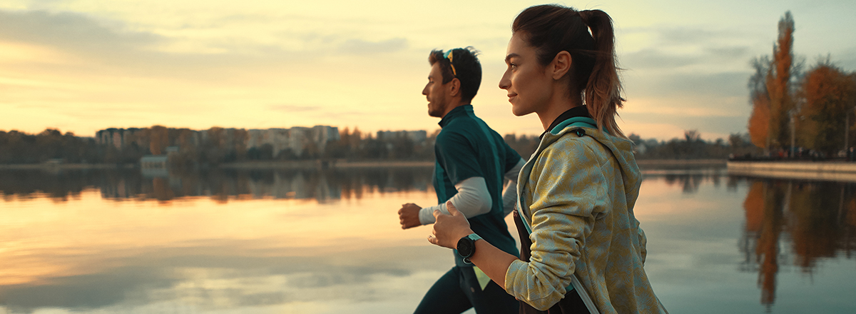 En ung man och en ung kvinna i träningskläder joggar. I bakgrunden syns en spegelblank sjö i gryningsljus samt natur och byggnader.
